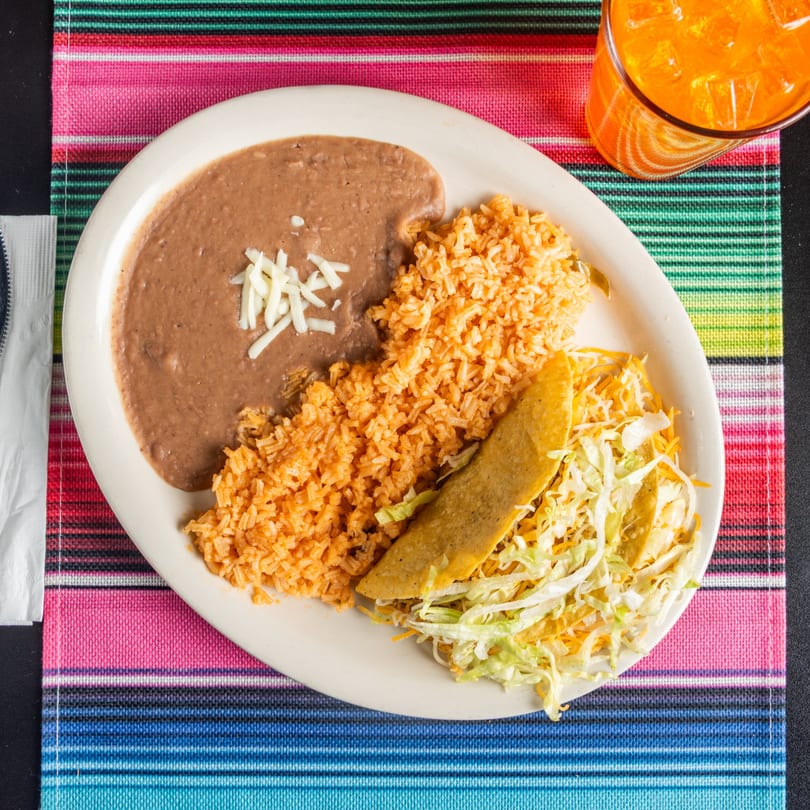 A Tacos 4 Ever plate with two tacos, rice, refried beans, and an orange drink.