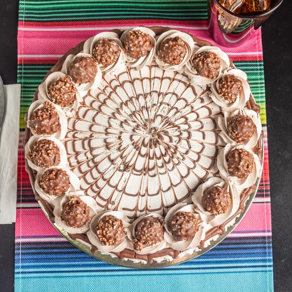 Round chocolate cake with whipped cream, chocolate drizzle, and Ferrero Rocher candies on a striped placemat.