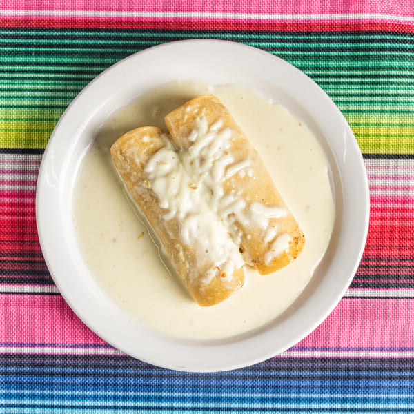 Two Tacos 4 Ever burritos covered in white cheese sauce on a white plate, on a colorful striped tablecloth.