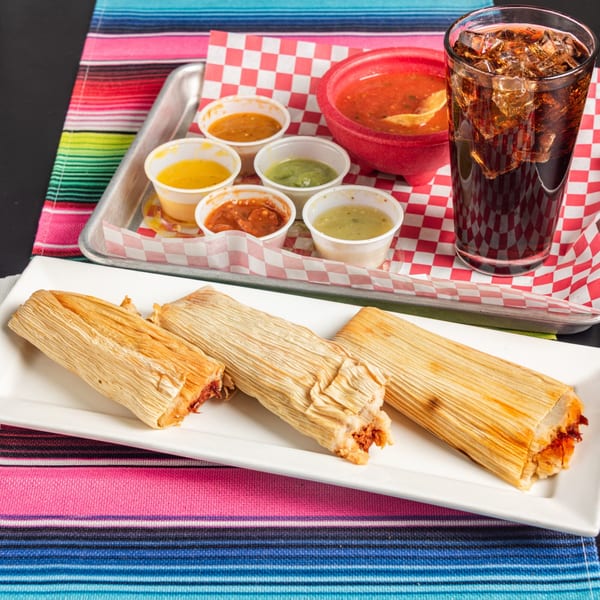 Three tamales on a white plate with a tray of various salsas, chips, and a soda.