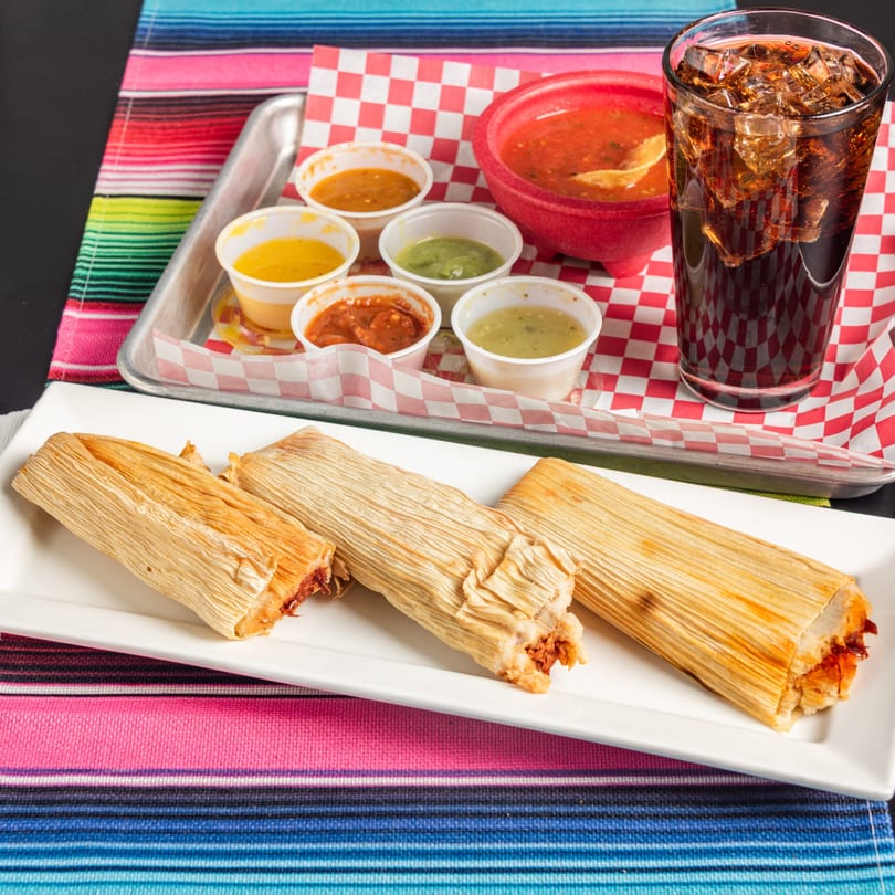 Three tamales on a white plate with a tray of various salsas, chips, and a soda.