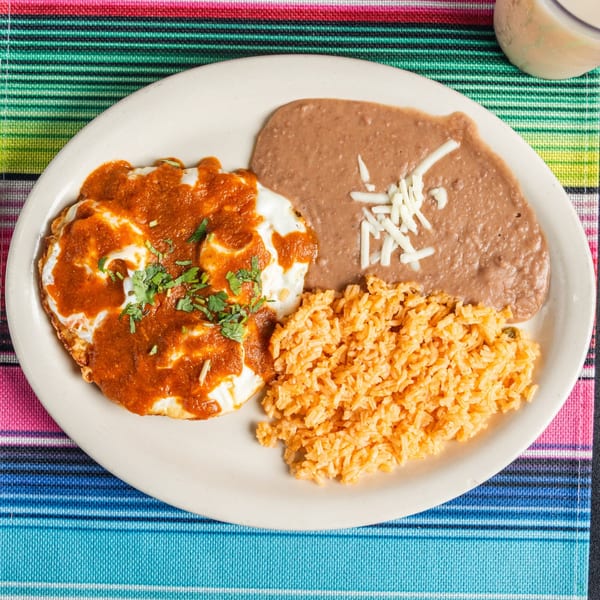 Huevos rancheros with red sauce, cilantro, refried beans, rice, and shredded cheese on a colorful placemat.