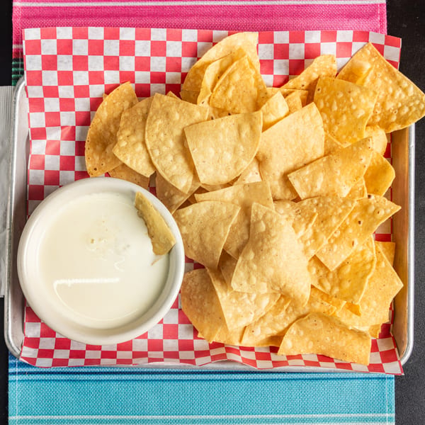A metal tray with a red and white checkered paper holds tortilla chips and a bowl of queso.