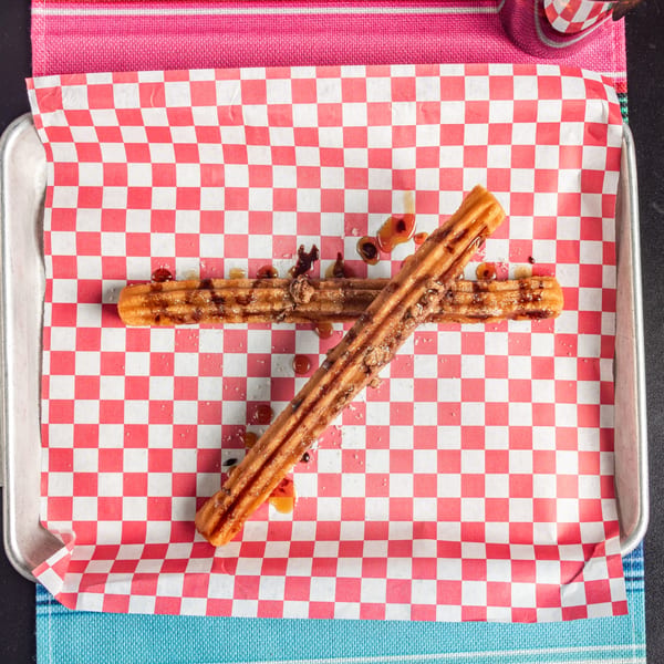 Two churros drizzled with caramel and chocolate sauce, on red and white checkered paper.
