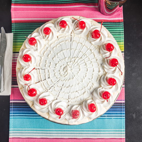 Overhead view of a whole tres leches cake with whipped cream and cherries on a colorful striped placemat.