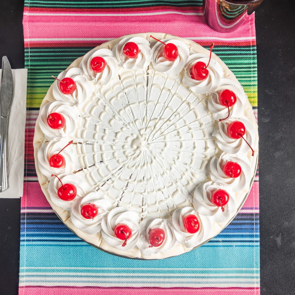 Overhead view of a whole tres leches cake with whipped cream and cherries on a colorful striped placemat.