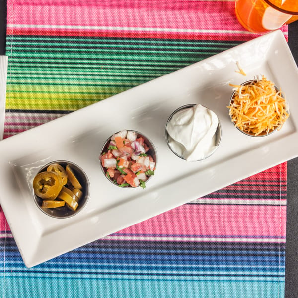 A white rectangular plate holds four small bowls of Mexican toppings: jalapeños, pico de gallo, sour cream, and shredded cheese.