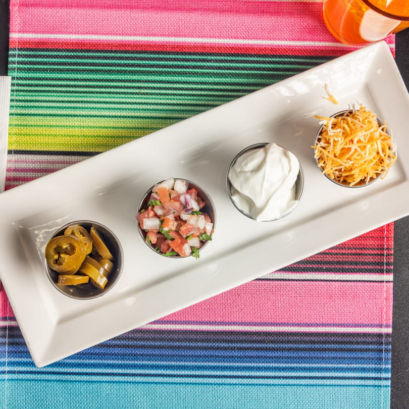 A white rectangular plate holds four small bowls of Mexican toppings: jalapeños, pico de gallo, sour cream, and shredded cheese.