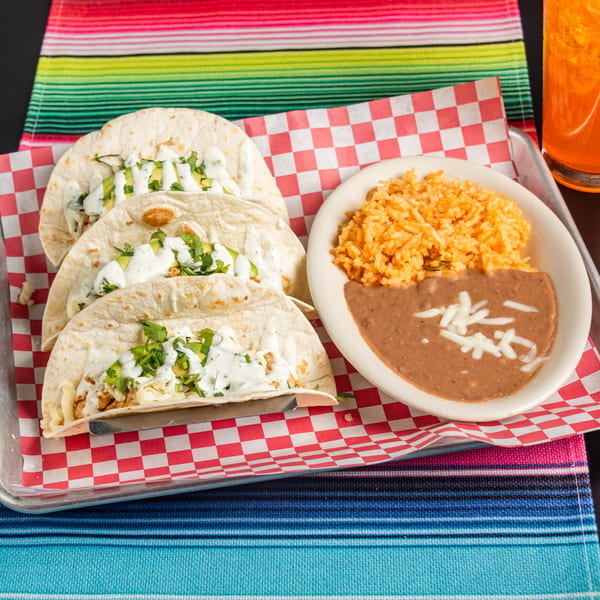 Three tacos with rice and beans, served on a metal tray with a colorful striped placemat.